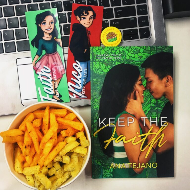 A flatlay of several items on a work desk: a small container of French fries, Bookmarks with drawn female and male characters, a book entitled "Keep the Faith by Ana Tejano" all on top of a laptop.
