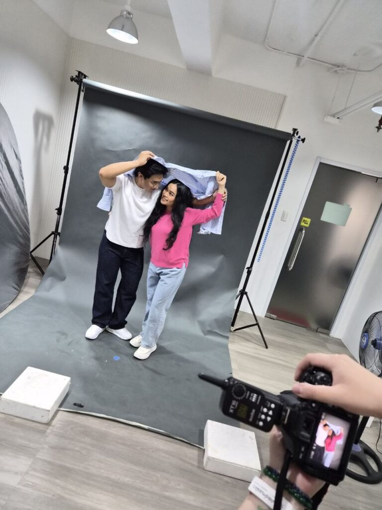 Man in white shirt and black pants posing with a woman in a pink knit top, holding a blue polo above them. A camera is on the lower right, taking their photo.